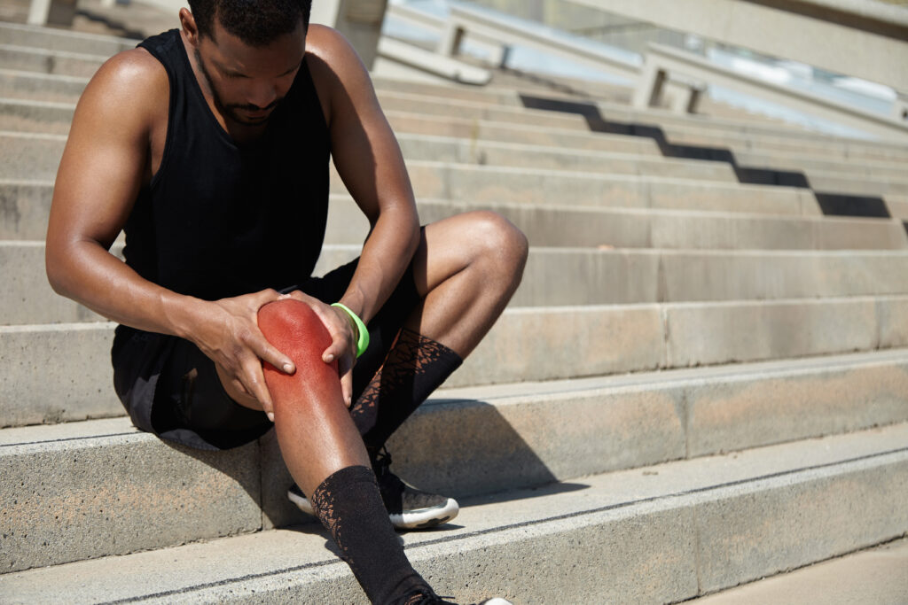 cropped portrait of dark skinned jogger in training socks and running shoes having sprain, clutching his injured knee, massaging it with both hands after intense workout in urban surroundings